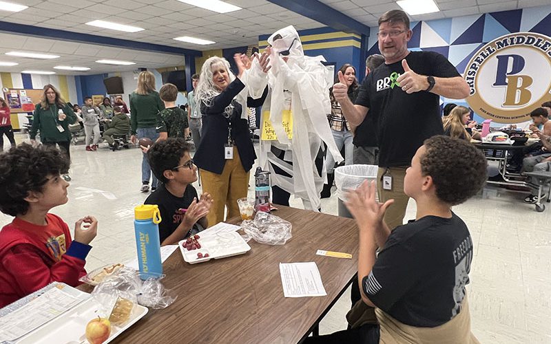A man all wrapped in toilet paper and wearing black sunglasses stands at a table of middle school students who give him thumbs up.