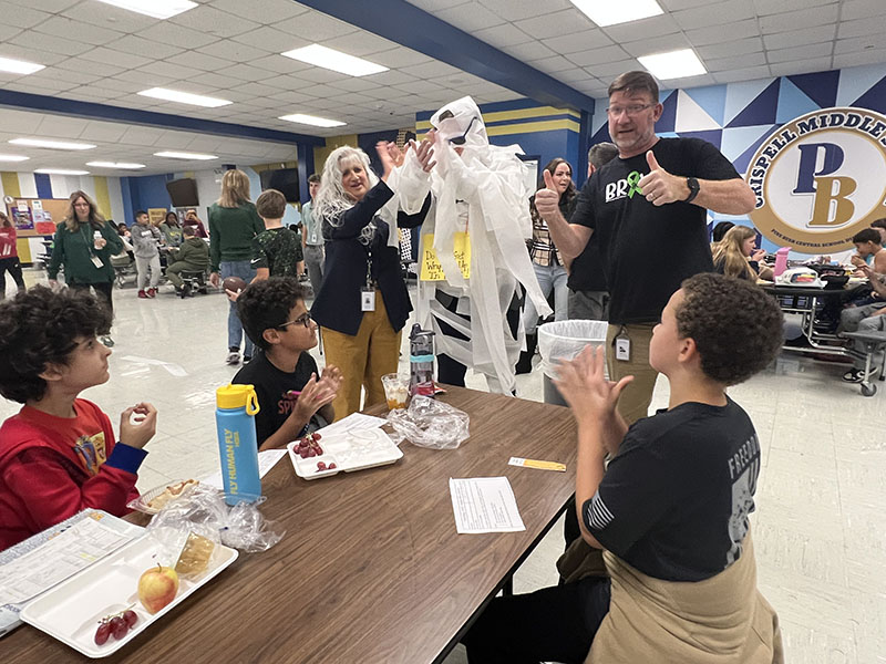 A man all wrapped in toilet paper and wearing black sunglasses stands at a table of middle school students who give him thumbs up.