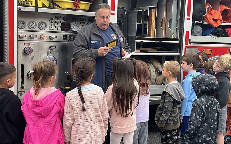 A man stands talking to a group of small elementary students. He is a fire fighter. Behind him is a fire engine.