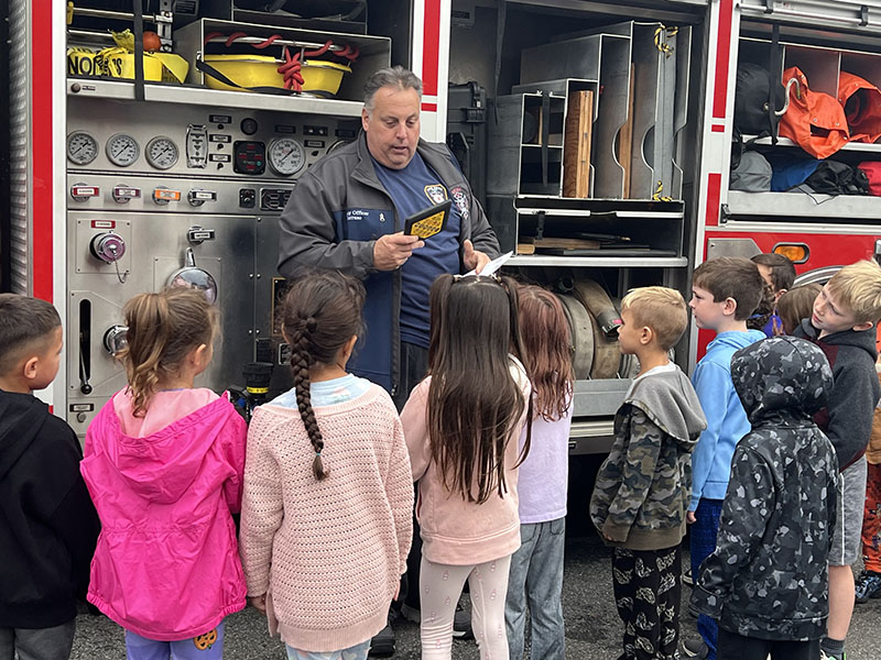 A man stands talking to a group of small elementary students. He is a fire fighter. Behind him is a fire engine.