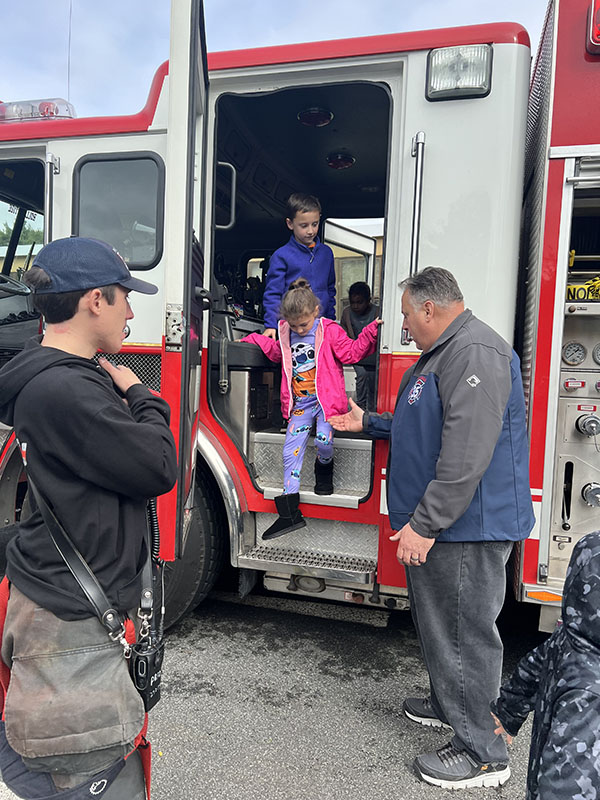 Small kids walk through a fire engine, with a firefighter on the outside to hold their hands coming down.