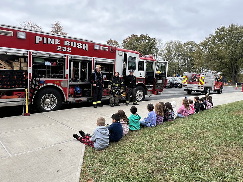 A group of small elementary children sit on grass and listen as a firefighter talks to them. Behind him is a large red fire engine that says Pine Bush FD.