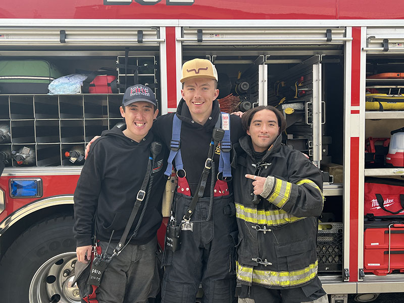 Three young firefighters stand together, arm in arm, and smile.