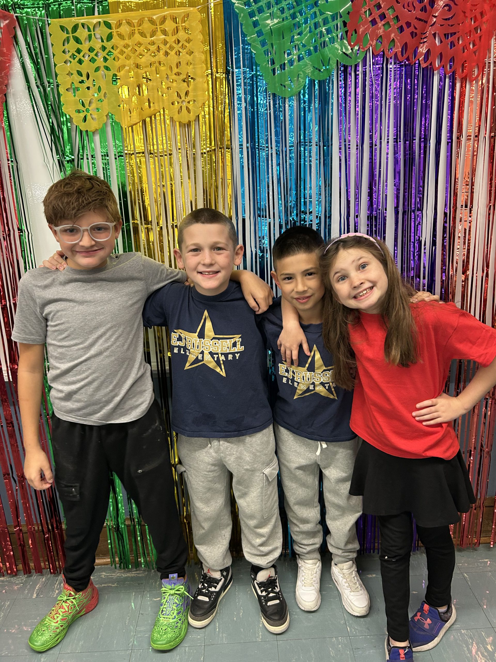 Shimmering colorful strands in the background. There are four third-graders standing arm-in-arm and smiling.