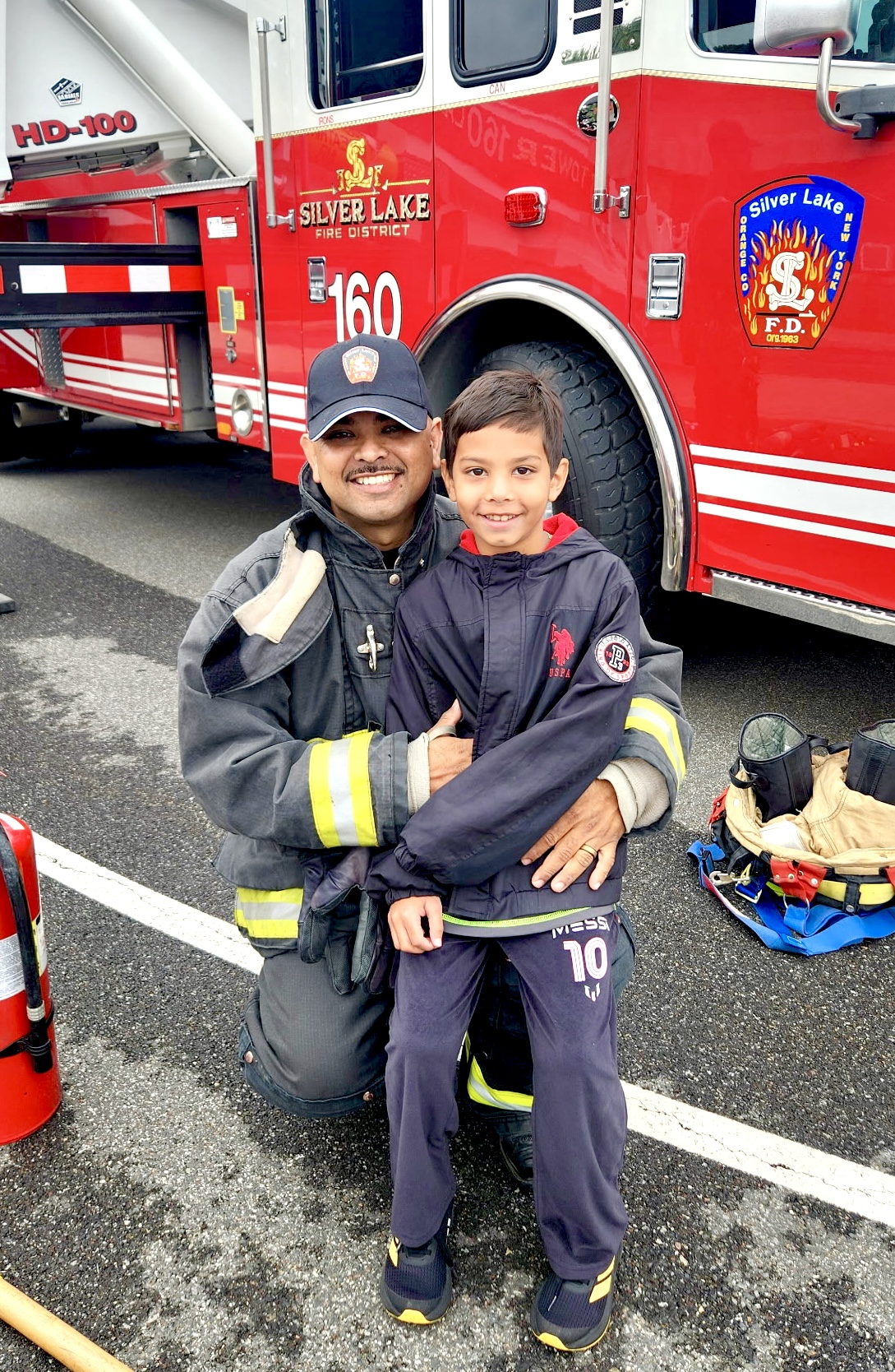 A man in a firefighter jacket smiles as a boy stands with him also smiling.