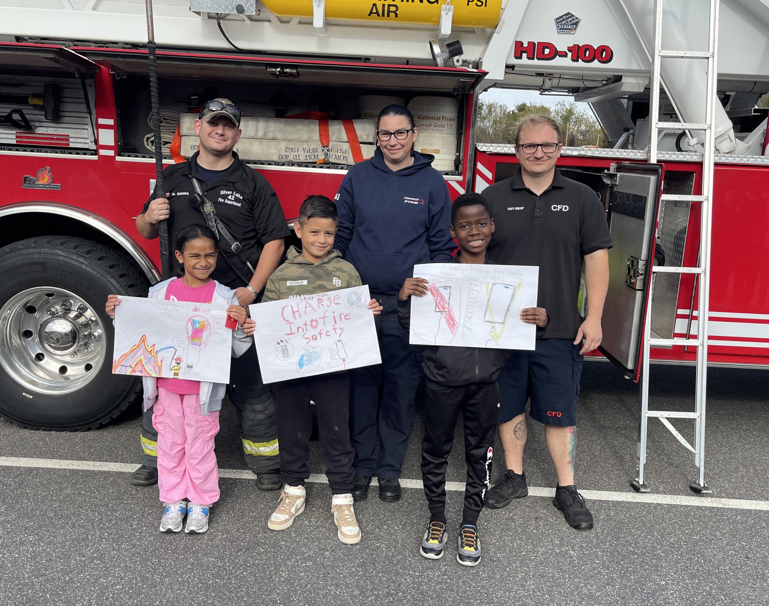 Three fire fighters stand behind three third grade students, who are holding posters they made about fire safety.