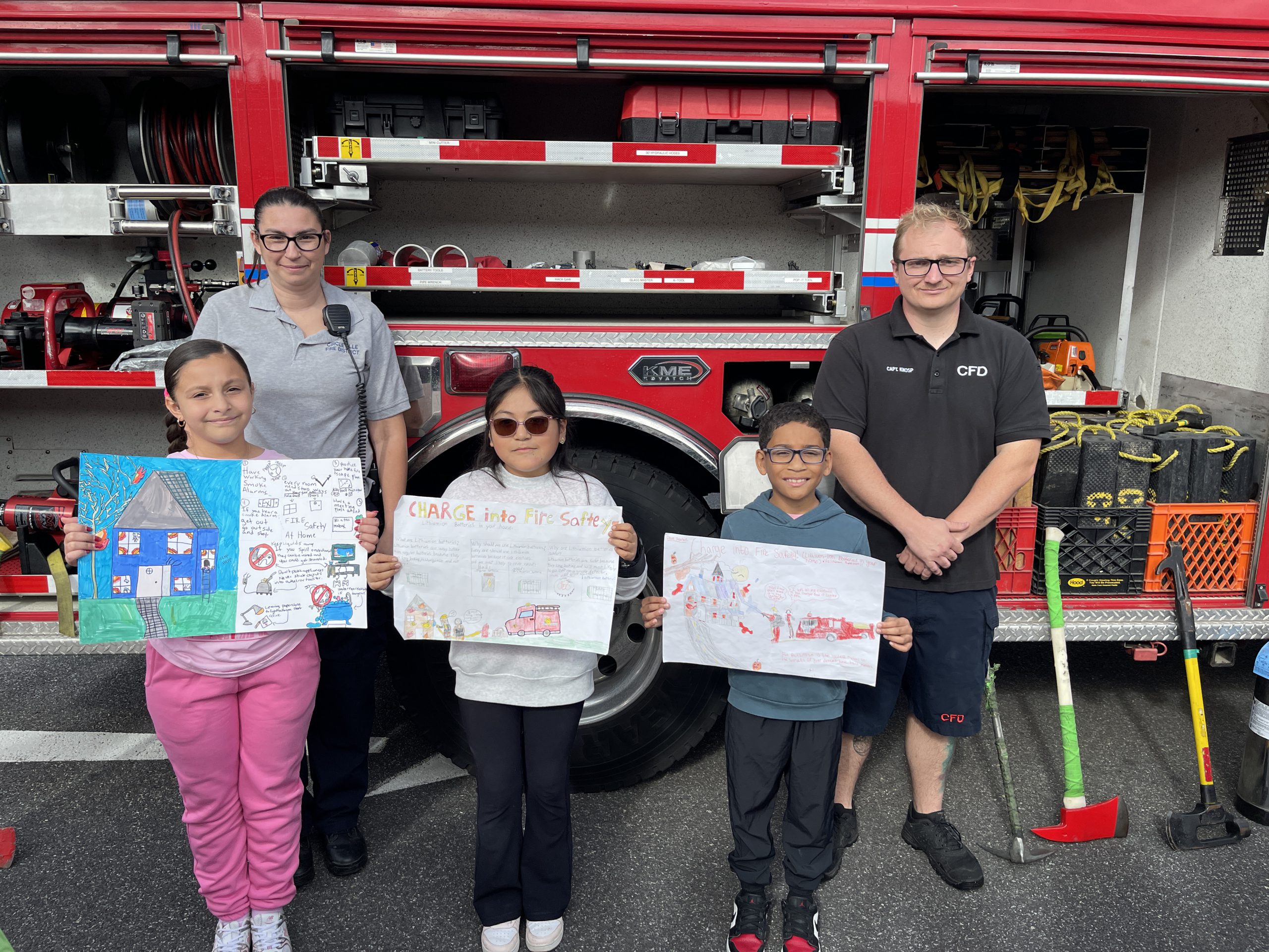 Three fourth graders stand in front of a red fire truck along with two firefighters.