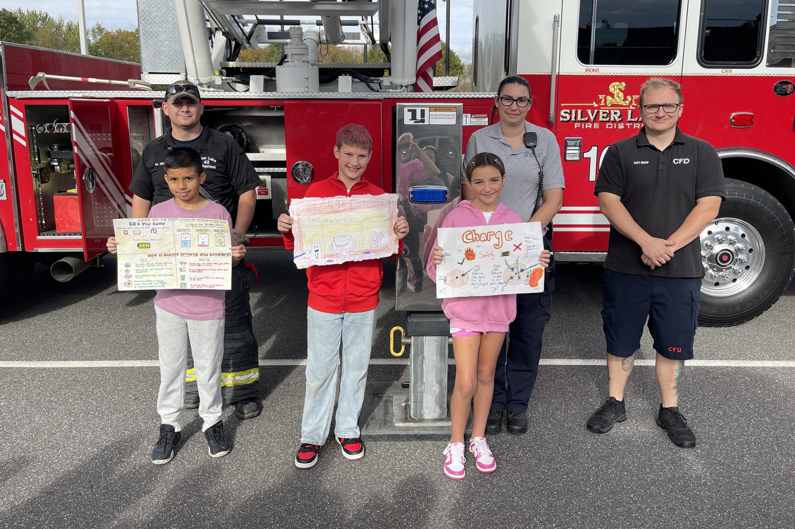 Three fifth grade students hold their posters standing in front of three firefighters. They are all standing in front of a fire engine.