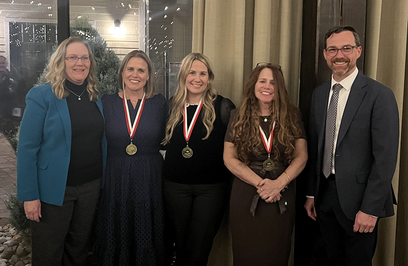 Five adults stand together and smile. The three women in the center all have medals around their necks after receiving awards of excellence.