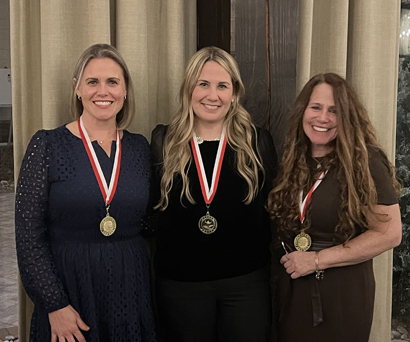 Three women stand together smiling. They have ribbons around their necks after winning awards.