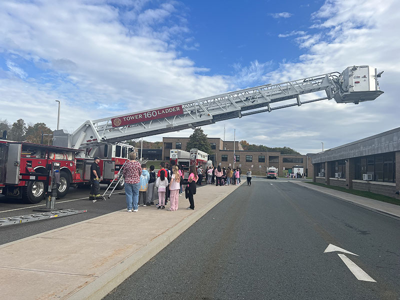 A very large ladder that says Tower Ladder extends over a group of kids listening to firefighters.