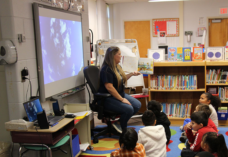 A woman with long blonde hair sits reading a book to a group of first grade students.