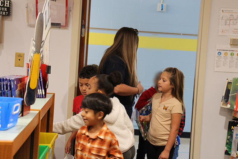 First grade students come into a library, putting their books into a slot.