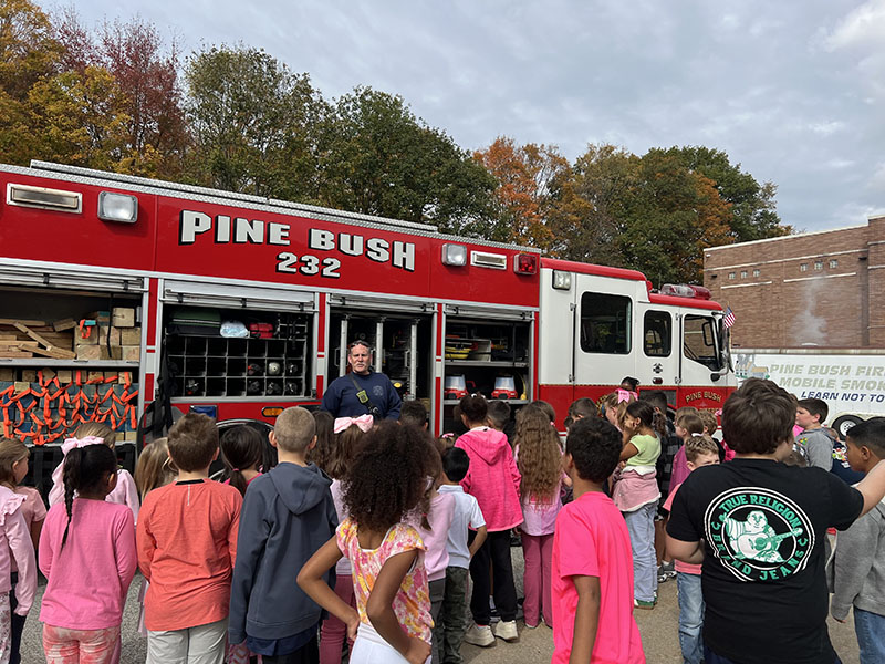 A red fire truck says Pine Bush FD on it. In front of it a firefighter talks to a group of older elementary students.