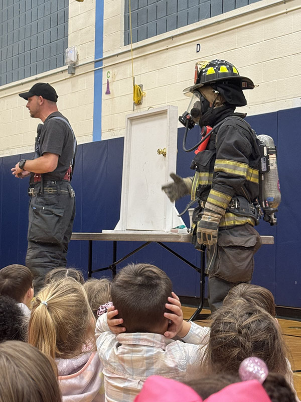 Two firefighters stand in front of a group of smaller elementary students who are sitting on the floor. One firefighter is in full gear.