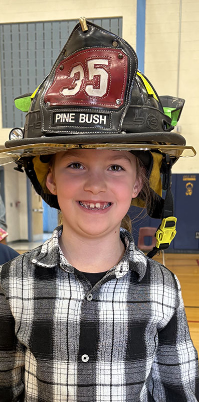 A younger elementary student smiles, wearing a large black firefighter helmet that says Pine Bush.