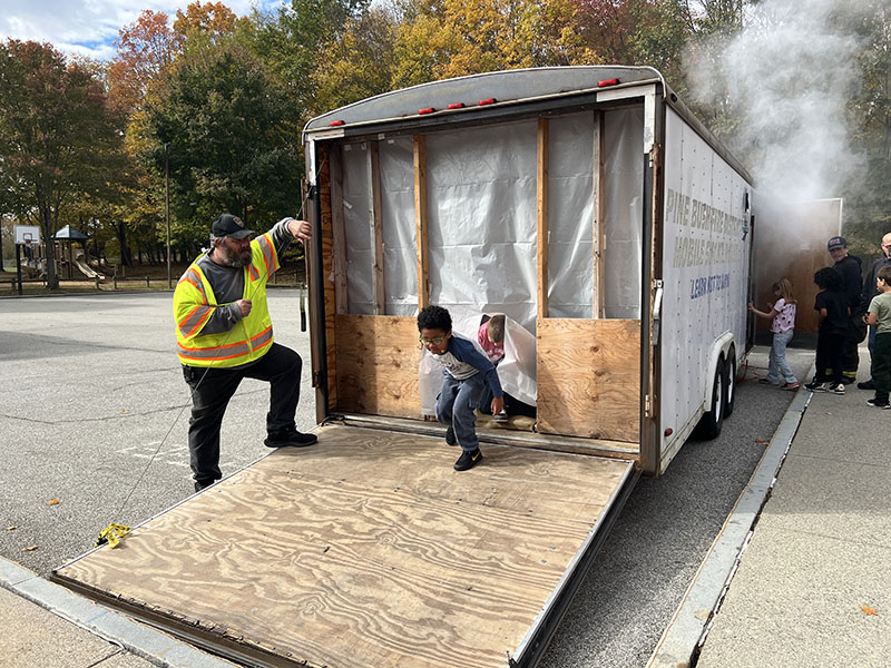 A gray trailer with a ramp coming out of it. A firefighter stands next to it ready to help the small elementary students who are coming out of it. There is a young boy coming down the ramp.