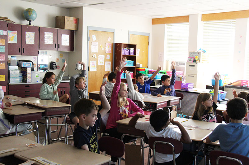 A classroom of third grade students, many with their hands up.