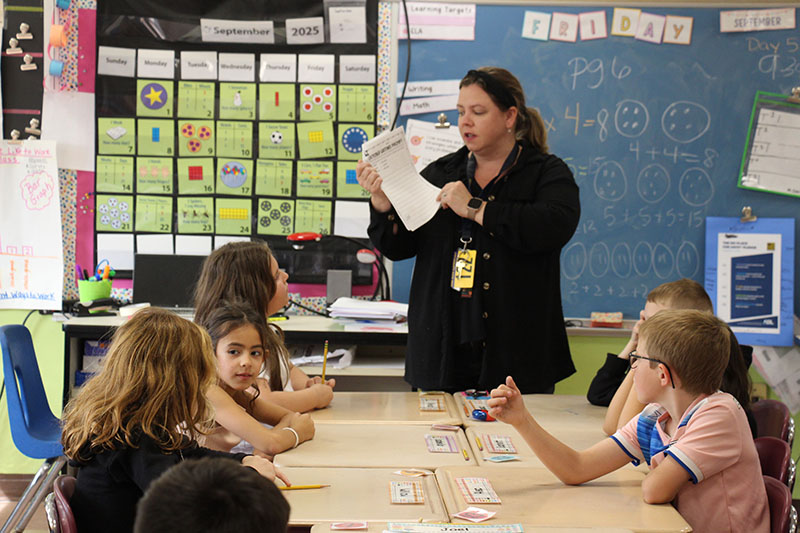 A woman stands in front of a table of third grade students. She is holding up a paper that says october Writing Prompts.