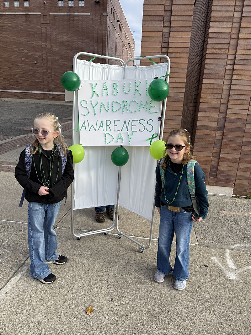 Two third grade girls, both with long blonde hair, wearing jeans and sunglasses, stand before a large sign that says Kabuki Syndrome Awareness Day.