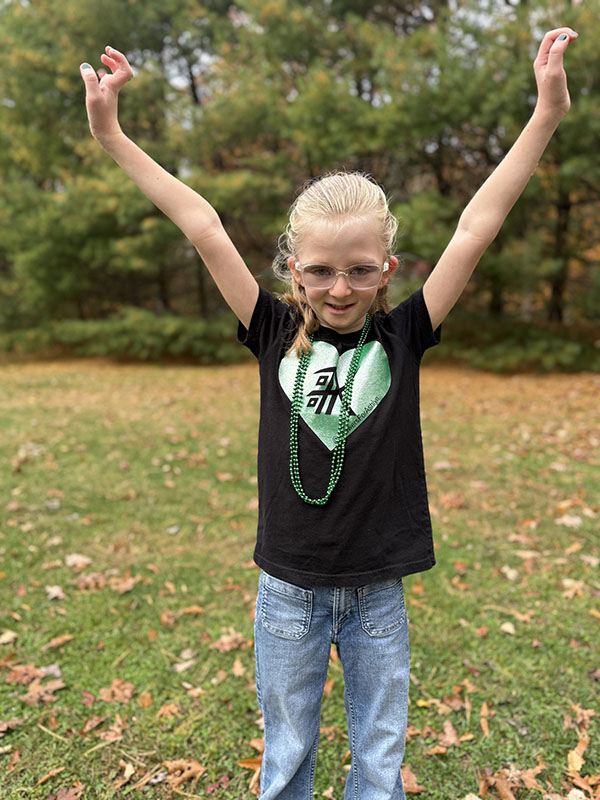 A third-grade girl with long blonde hair stands with her hands up. She is wearing a black shirt with a green heart on it.