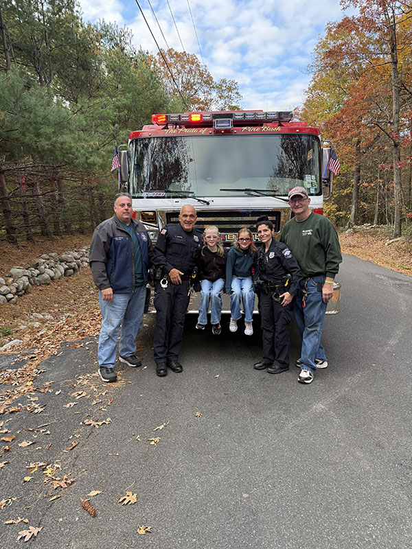 Two third-grade girls sit on the front of a large fire engine. Next to them are four adults.