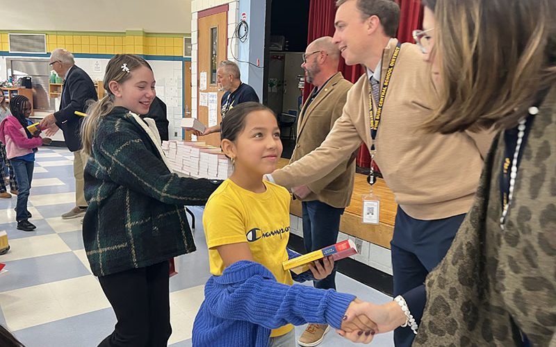 A third-grade girl in a yellow shirt shakes hands with a woman after receiving a red dictionary.