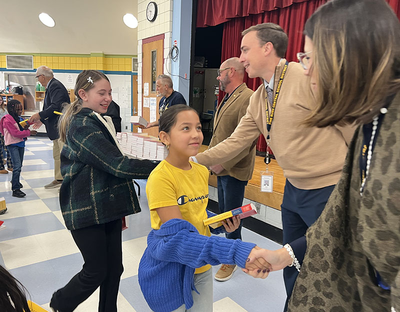 A third-grade boy in a yellow shirt shakes hands with a woman after receiving a red dictionary.