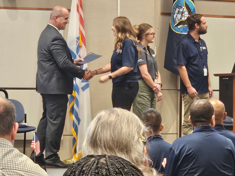 A woman shakes hands with a man who is holding a diploma in his other hand. Others are around watching.