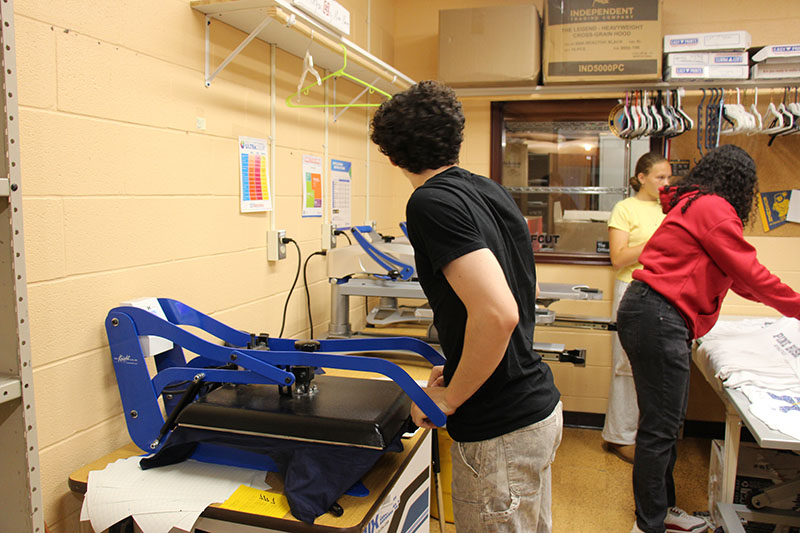 High school kids working in the school store. One is using a press to make tshirts and two others are laying out the newly made shirts.