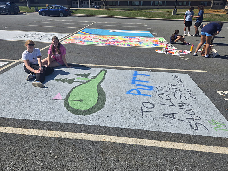 A parking lot with high school students painting the spaces. The painted spot has a putting green on it.