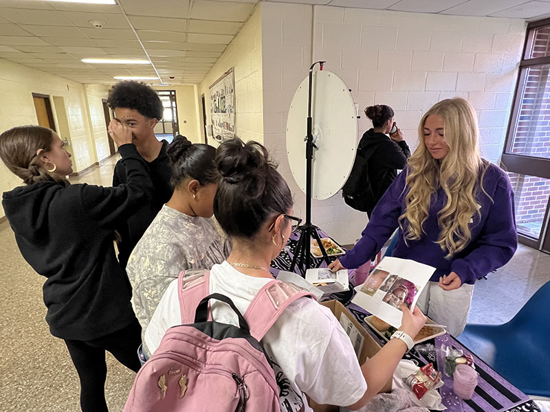 a high school girl with long blonde hair hands a piece of paper to another girl standing at the table with her. Another girl is also there. Next to them is a  high school boy getting his face painted.