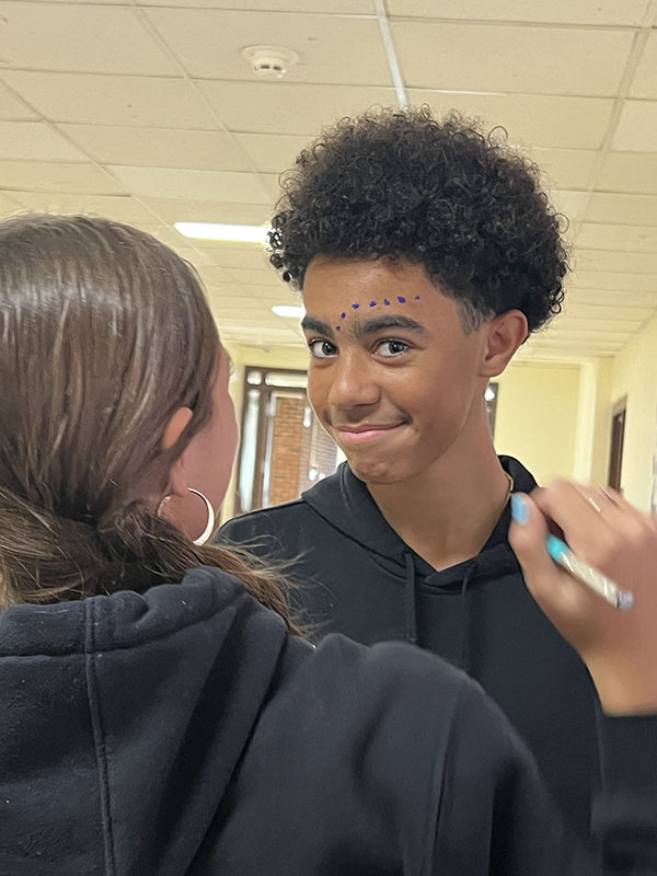 A high school student smiles. His face has purple dots painted around his eyes. A high school girl stands in front of him with a paintbrush.