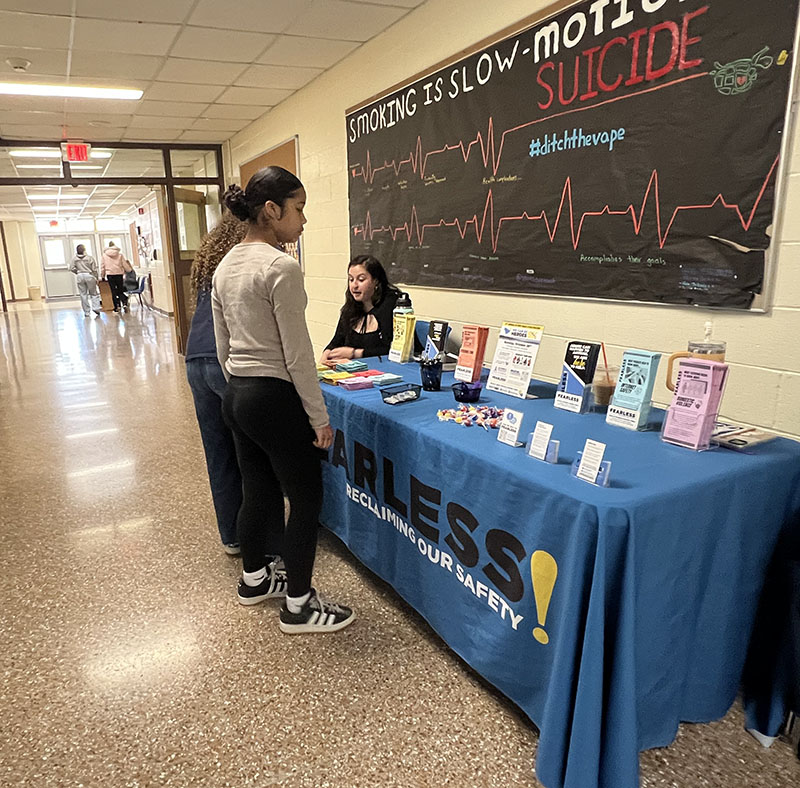 A table with a blue tablecloth that says Fearless! on it is in a hallway. There is literature on the table and two high school students standing there looking through it. A woman is sitting at the table.