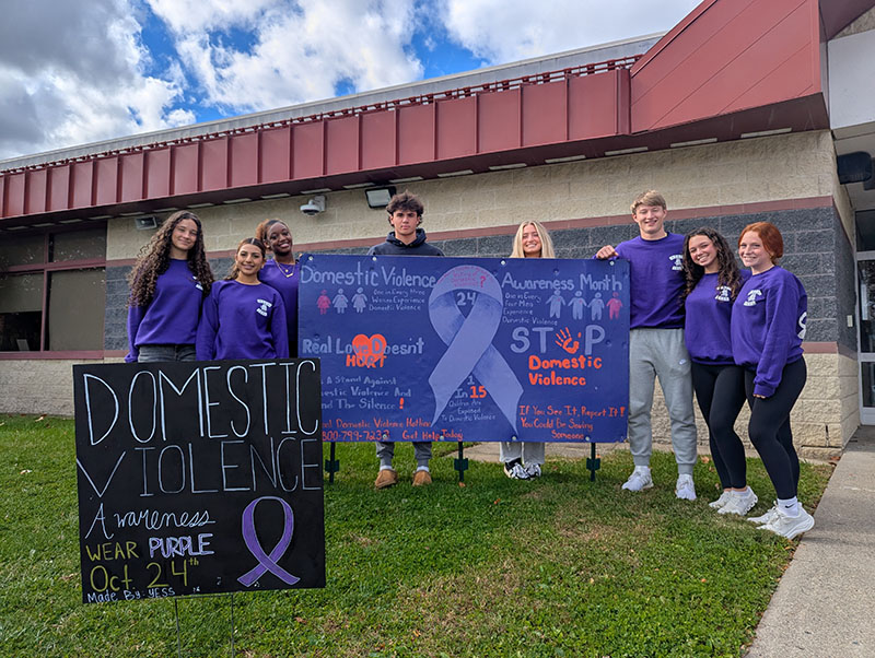A group of eight high school students stand around a large purple sign that says Domestic Violence Awareness Month Stop Domestic Violence Real Love Doesn't Hurt.