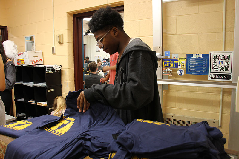 A high school kid folds and sorts through a large stack of navy blue tshirts with gold writing.