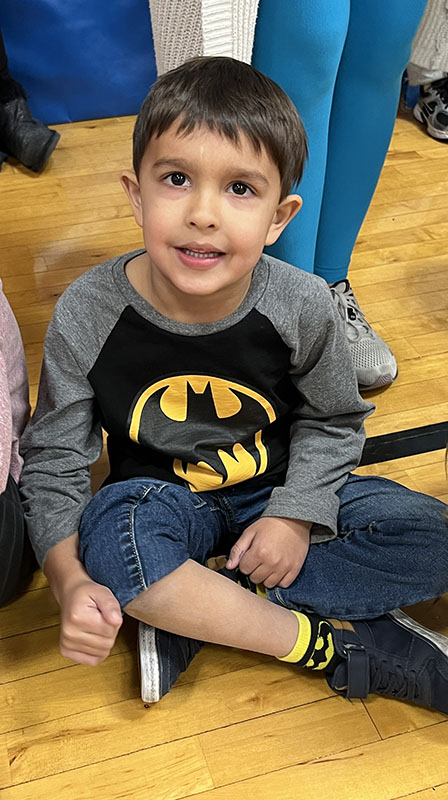 An elementary age kid sits on a gymnasium floor. He is wearing a batman shirt.