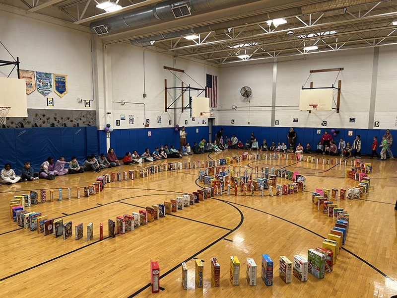 A large gymnasium with kids all around the sides. In the center is a large line of cereal boxes set up as dominoes.