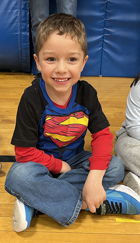 An elementary age kid sits on a gymnasium floor. He is wearing a superman shirt.