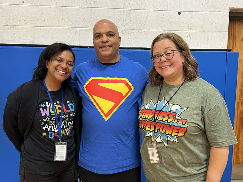 Three teachers stand arm in arm and smile. They are wearing superhero shirts.