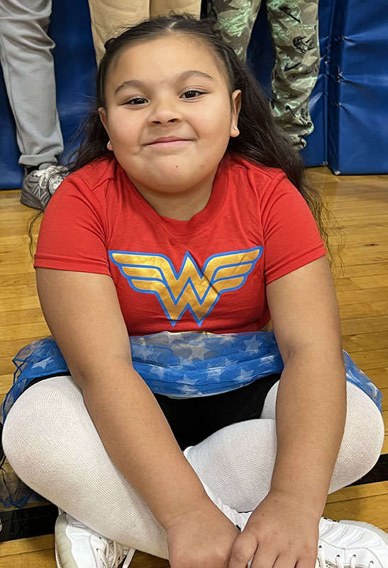 An elementary age kid sits on a gymnasium floor. She is wearing a Wonder woman shirt.