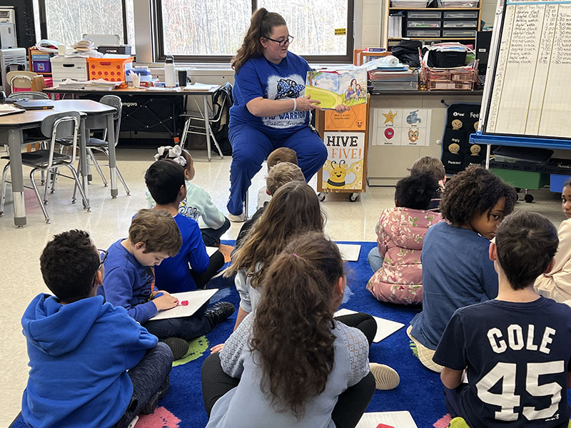 A woman in blue scrubs sits on a chair in front of a class and reads a book.