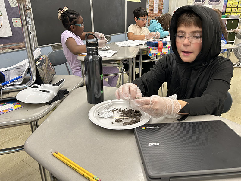 An elementary age boy wearing a black sweatshirt works on a science project at his desk.
