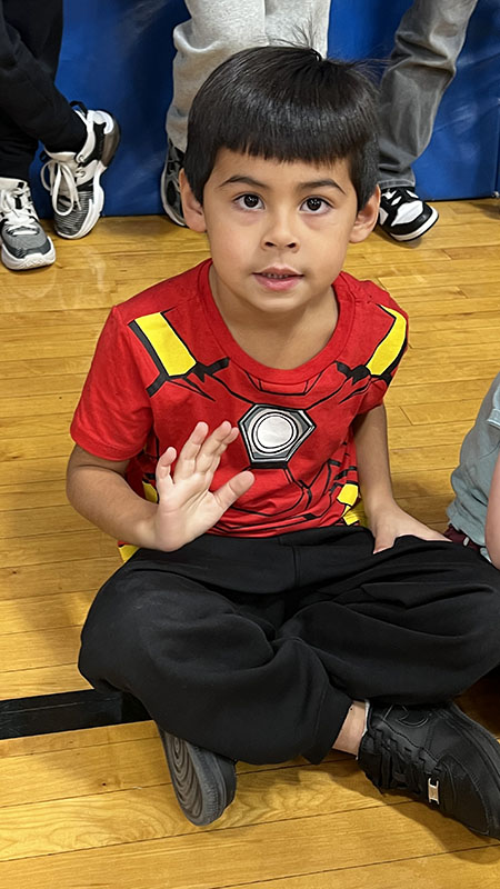 An elementary age kid sits on a gymnasium floor. He is wearing a spiderman shirt and waving.