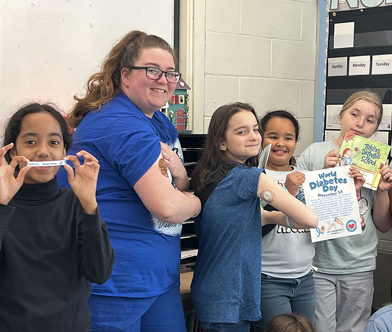 A woman in blue scrubs smiles and shows her diabetes monitor on her arm. She is surrounded by fifth-grade girls. One is flexing her arm showing her monitor too.