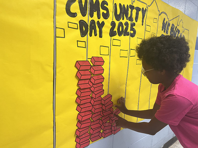 A middle school student takes a piece of red paper that is in the shape of a brick and adds it to the mural on the wall.