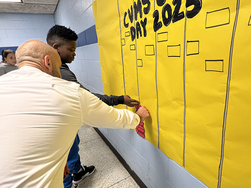 A man leans in to help a middle school student afix a red piece of paper to a yellow mural.