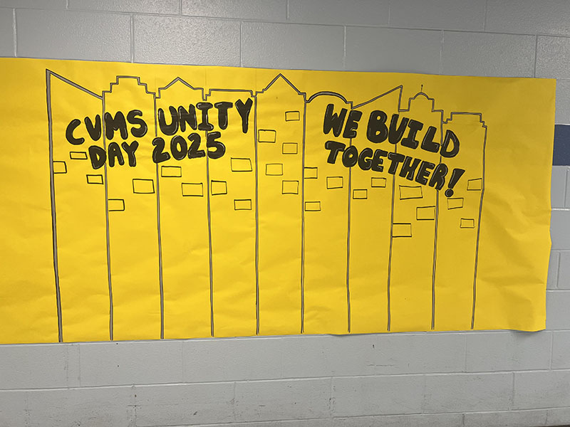 A very large mural of yellow paper is on a wall. It says CVMS Unity Day 2025. We Build Together! The outline is of tall buildings.