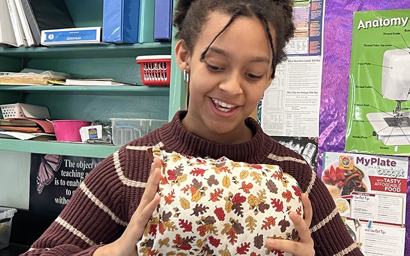 A girl smiles as she holds up her small rectangular pillow. She looks down at it. The pillow has autumn leaves on it.