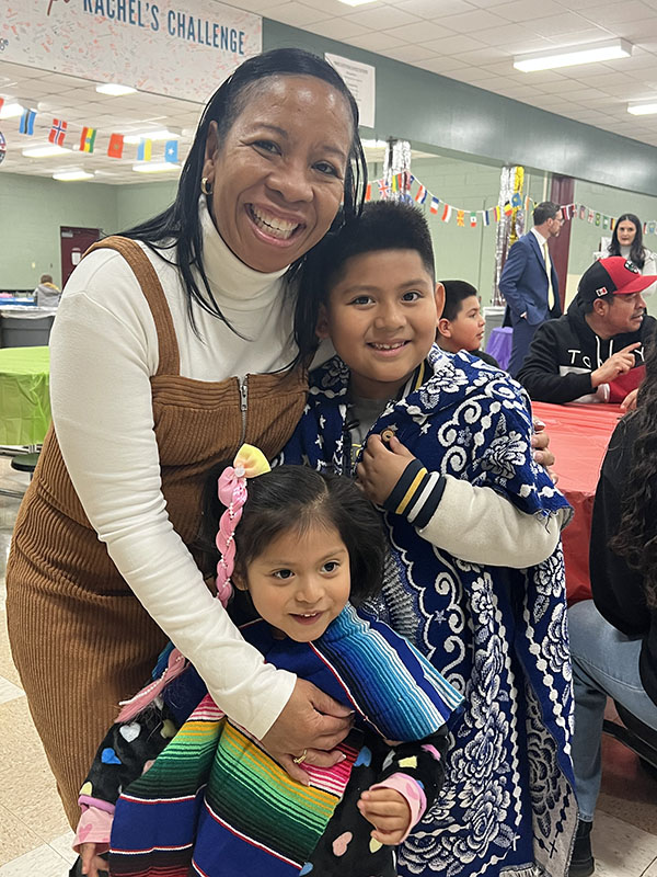 A woman wearing a white sweater and brown jumper hugs two young children dressed in clothes from their home country.